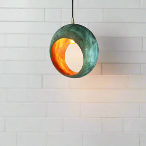 A spherical pendant light with a weathered greenish-blue exterior and a warm orange interior, hanging against a white tiled wall