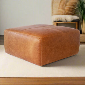 Handcrafted square tan leather ottoman in a cozy living room with a neutral rug, wooden furniture, and natural decorative elements.