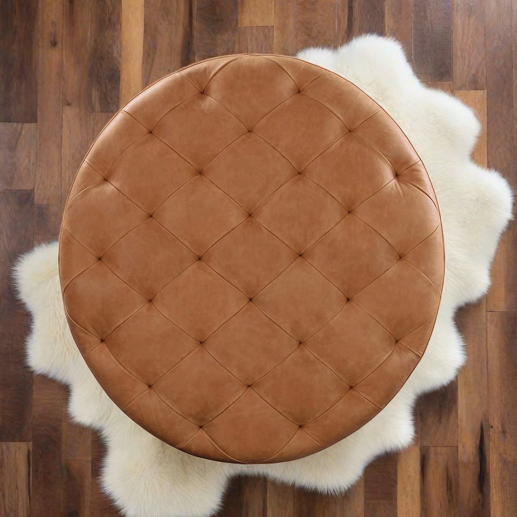 An overhead view of a round, tufted leather ottoman in a warm caramel brown color, positioned on a fluffy white sheepskin rug atop a wooden floor.