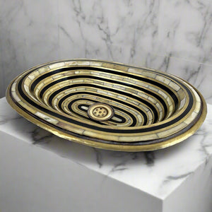 A close-up of a decorative bathroom sink with an intricate black and gold geometric pattern. The sink has a brass drain and is paired with a modern brass faucet. The sink is mounted on a white marble countertop, adding a luxurious touch to the bathroom decor