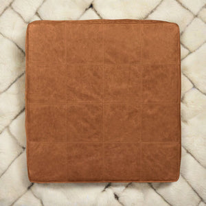 Top view of a handcrafted square tan leather ottoman showcasing its detailed stitching and high-quality leather finish on a light patterned rug.