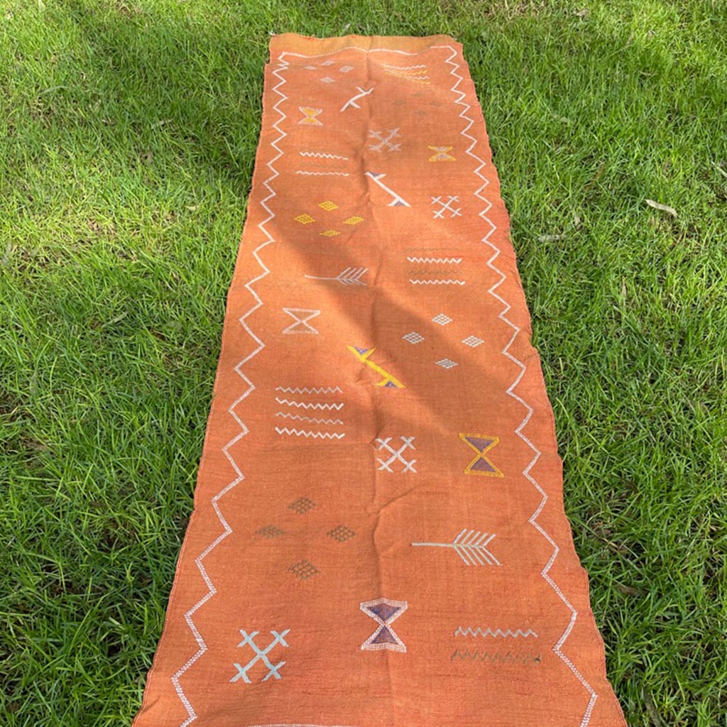 Top-down view of a long terracotta Moroccan runner rug with traditional Berber patterns and a zigzag border, laid flat on a grassy lawn.