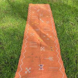 Top-down view of a long terracotta Moroccan runner rug with traditional Berber patterns and a zigzag border, laid flat on a grassy lawn.