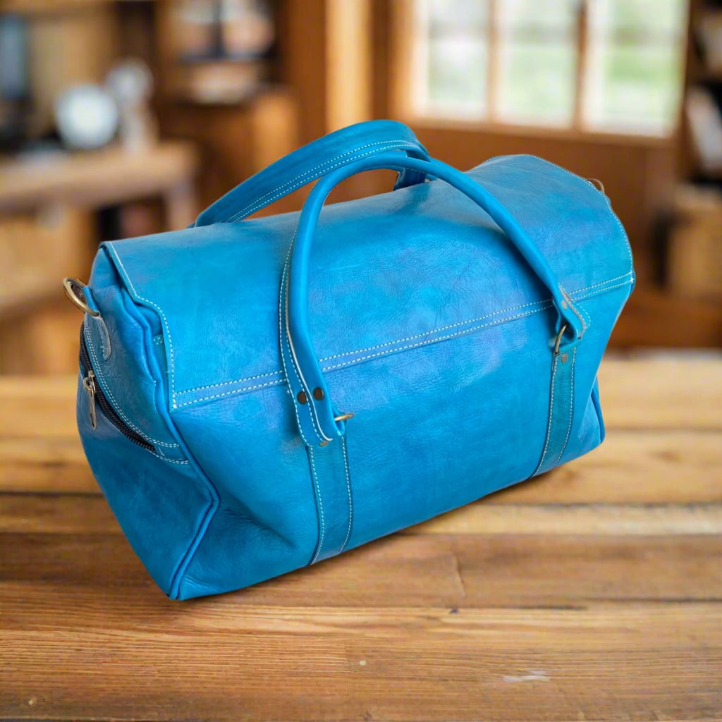 A sleek blue leather duffle bag with minimalist stitching, sturdy handles, and a zippered side pocket, set on a rustic wooden table with a softly lit background.