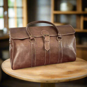 A vintage brown leather duffle bag crafted from premium leather, featuring a front buckle closure, dual stitched handles, and a timeless, minimalist design. The bag includes reinforced stitching for durability, a spacious structure, and subtle detailing that enhances its classic appeal. It is placed on a round wooden table in a warm, softly lit interior, highlighting its rich brown tone and elegant craftsmanship