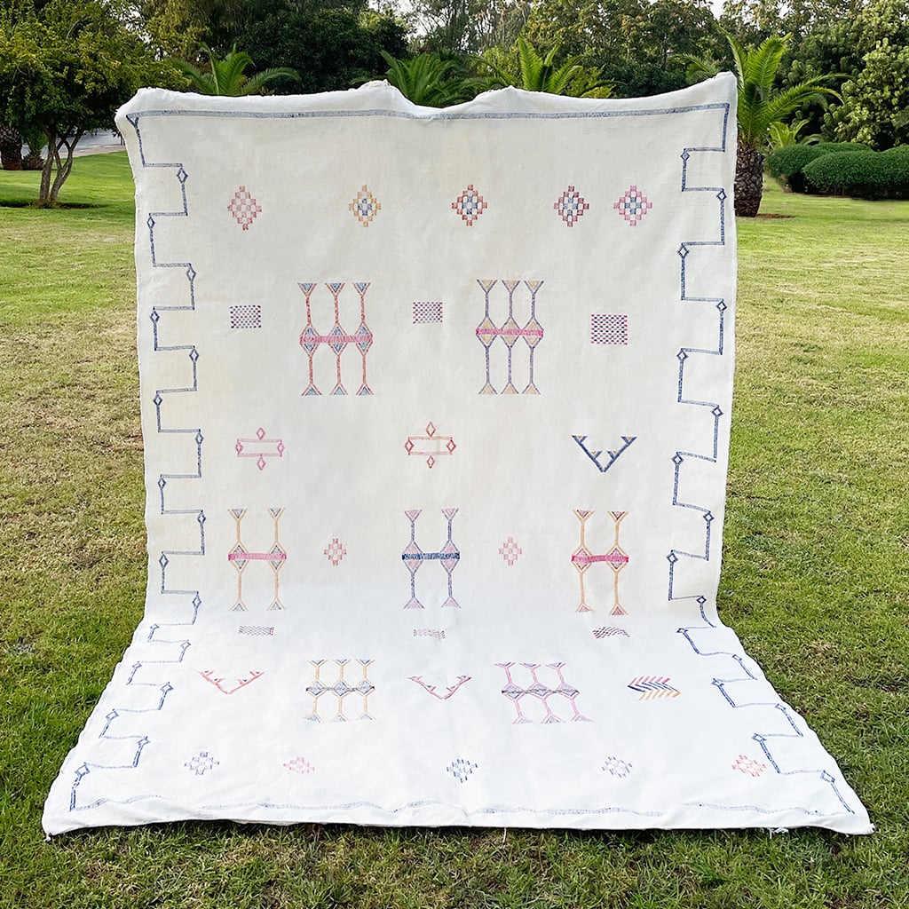 White Moroccan sabra cactus silk rug with colorful hand-embroidered Berber motifs and tribal symbols, displayed upright on green grass