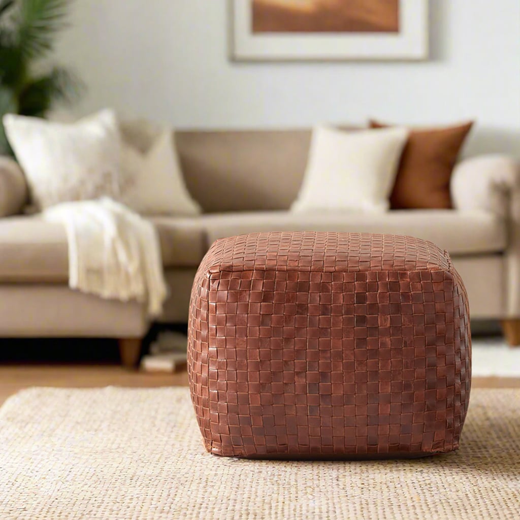 A brown woven leather pouf sits on a textured beige rug in front of a neutral-toned sofa with white and brown pillows. A framed artwork hangs above the sofa.