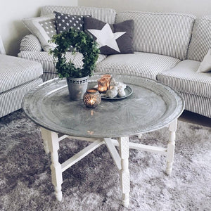 A silver Moroccan tray table with a white base, topped with decorative candles and a plant, in a cozy living room with striped couches