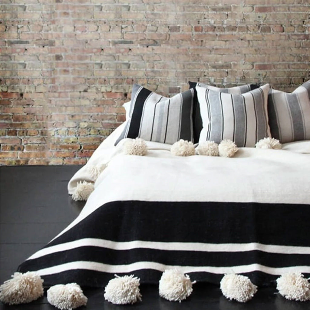 Moroccan Pon pom blanket on top of a bed with striped pillows against a brick wall in a living space - Moroccan Interior