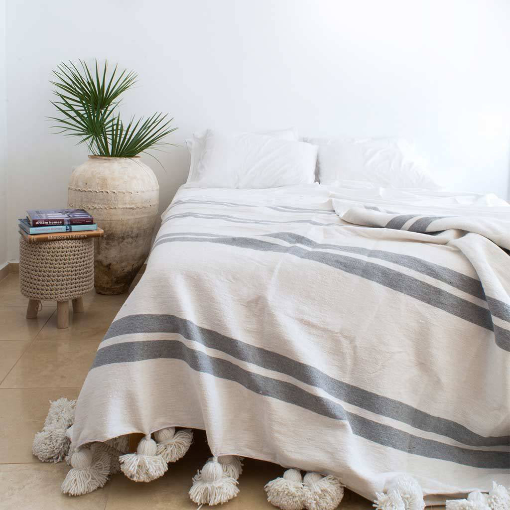 A cozy bedroom with a Moroccan cotton pom poms blanket in gray striped pattern draped over a bed. The blanket features soft white pom-poms along the edges, complementing the minimalist decor with a neutral color palette and a potted plant in the background