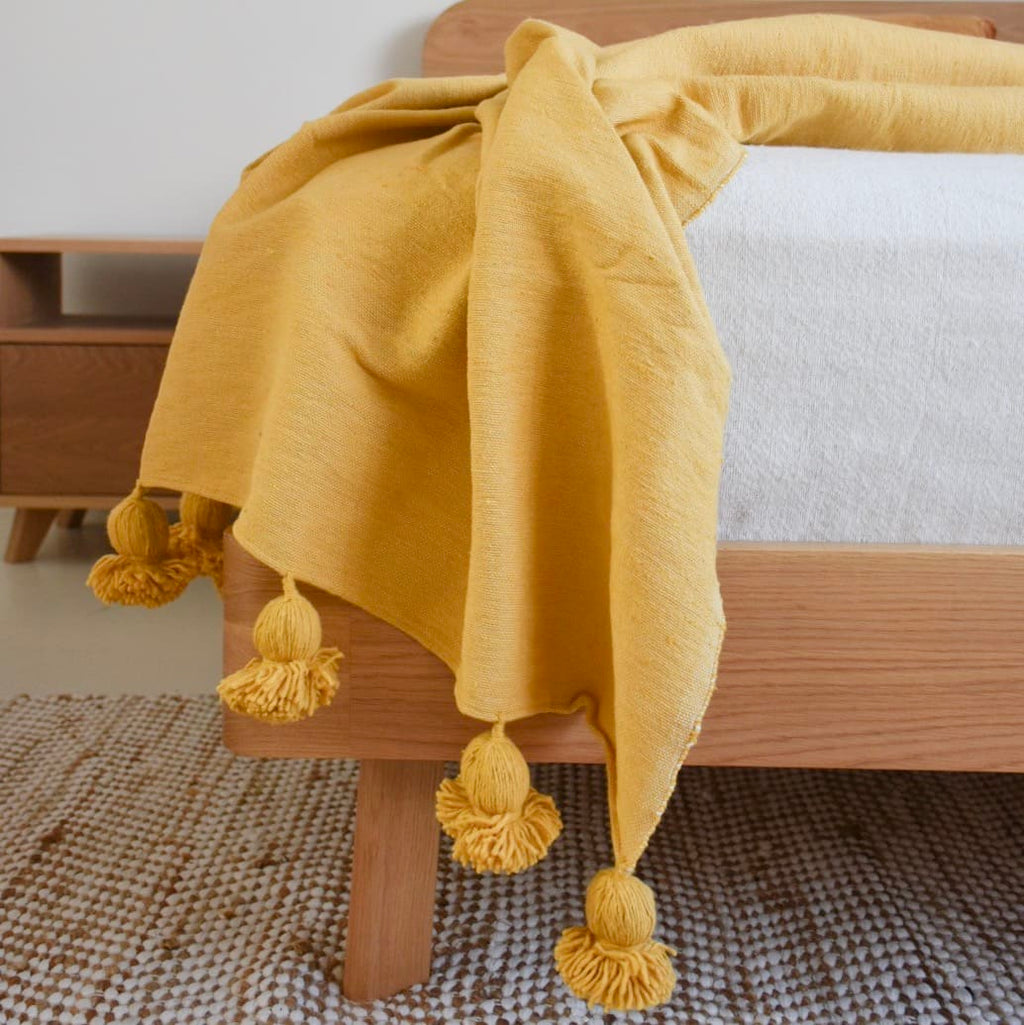 Yellow Moroccan cotton pom pom blanket draped casually over the edge of a wooden bed, with large pom poms hanging off the sides. The soft, handwoven texture and rich yellow color are showcased against a neutral room setting