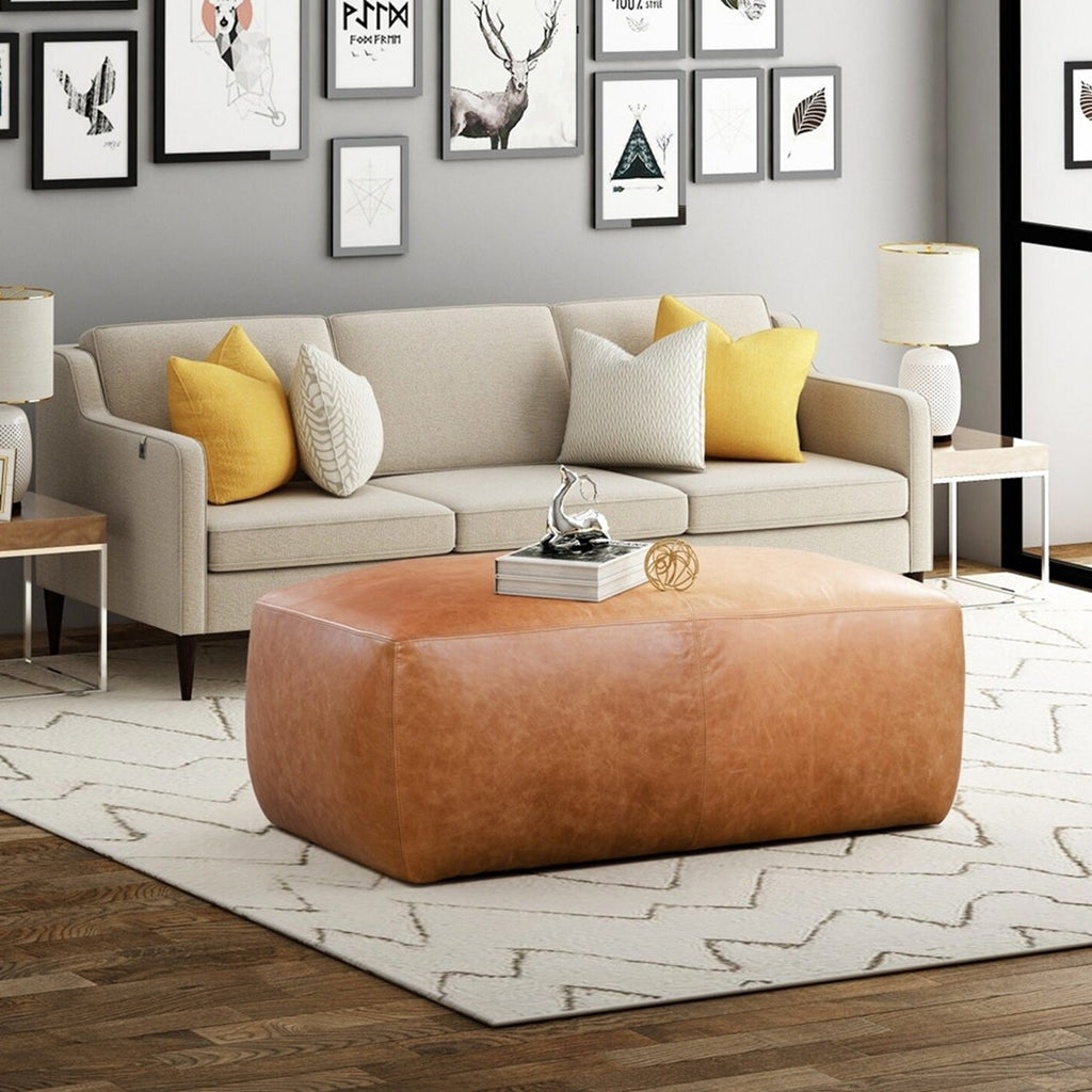 Stylish Moroccan-inspired living space: Plush 3-seat sofas, pillows, picture frames on light gray wall, central tan leather pouf - Moroccan Interior