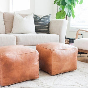 Set of two Moroccan leather poufs on wool rug in living room with furniture, big plant, and window - Moroccan Interior
